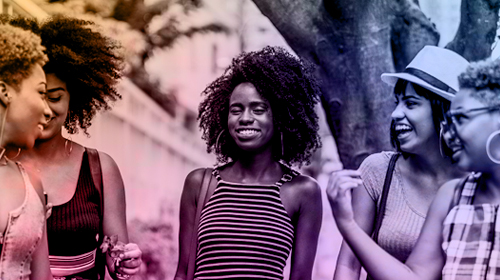 Na imagem vemos um grupo de cinco mulheres em semicírculo, sorrindo umas para as outras.