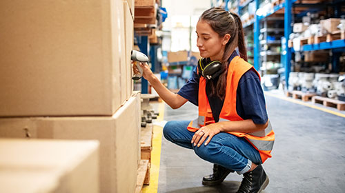 Em um ambiente de estoque de materiais, uma mulher está agachada, com um leitor de código de barras na mão conferindo caixas que estão apoiadas no chão.