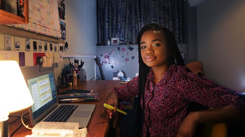 A imagem é uma foto. Em um quarto, que também é escritório, uma mulher negra, de tranças e camisa florida de mangas longas, está sentada em uma cadeira, segurando uma caneta marca-texto e olhando para a câmera. Em frente a ela está uma mesa de madeira, com um notebook aberto, um abajur e outros materiais, como canetas e papéis coloridos.
