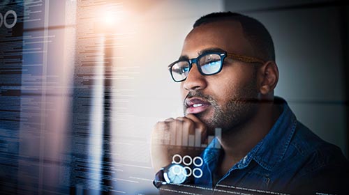 A imagem é uma foto. Um homem negro, de cabelos curtos, óculos, barba, camisa social jeans e relógio, está com a mão apoiada no queixo. Sua imagem está refletida em uma tela de computador, onde também estão informações escritas.