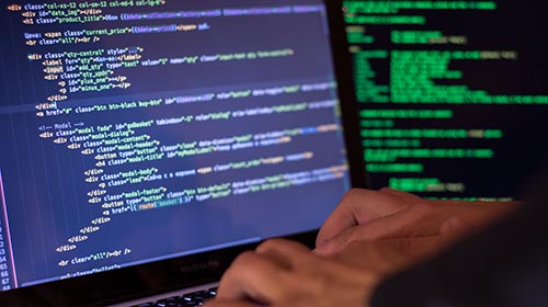 A imagem é uma foto. As mãos de um homem branco digitam no teclado de um computador. Na tela estão códigos de programação. Uma tela de computador, ao fundo, também exibe códigos.