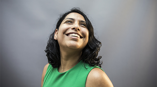 A imagem é uma foto. Uma mulher latina, de cabelos pretos, na altura dos ombros, sorri e olha pra cima