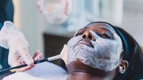 A imagem é uma foto de uma mulher negra deitada com os olhos fechados. Em seu rosto, um creme branco está sendo espalhado com o auxílio de um pincel manipulado por uma pessoa utilizando luva branca.