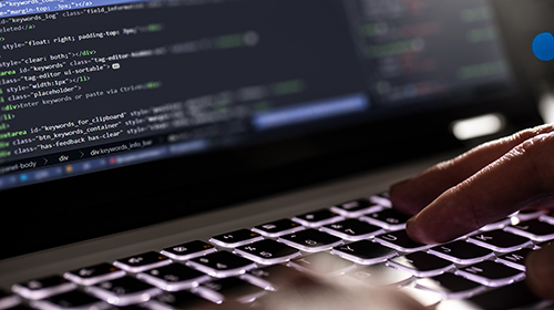 A imagem é uma foto. O foco está em um computador e, na tela, aparecem vários códigos de programação. O teclado do computador é luminoso. Sobre ele, um par de mãos está digitando