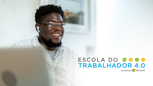 A imagem é uma foto. Um homem negro, de cabelos curtos e de óculos, está em um ambiente de escritório. Ele está sentado, usa fones de ouvido e tem duas telas de computador à sua frente.