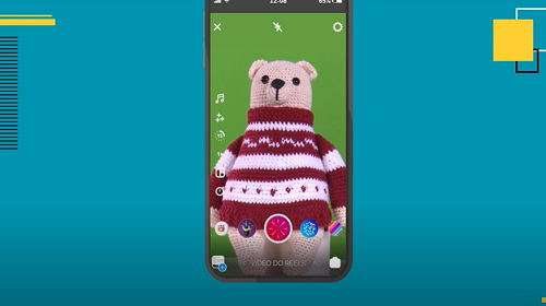 A imagem é uma foto com intervenções gráficas. Em um fundo azul está aplicado um celular preto. Dentro do celular, está um fundo verde com um urso feito com a técnica de crochê japonês, amigurumi. No canto esquerdo da tela do celular aparecem ícones representando música, brilho, tempo e velocidade.