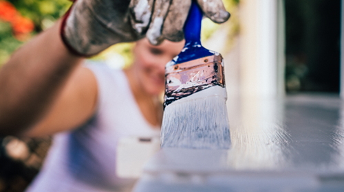 A imagem é uma foto. Um homem branco, calvo, usando óculos de proteção e camiseta azul, está em uma sala. Ele pressiona uma furadeira contra uma porta de ferro preta.