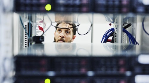 A imagem é uma foto. O rosto de um homem negro, de cabelos curtos e barba, está entre dispositivos de hardware, com luzes e fios pendurados