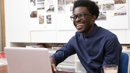 A imagem é uma foto. Em um ambiente de escritório, um homem negro, com óculos de armação preta e camisa azul, usa um notebook que está em cima de uma mesa na frente dele. Ao lado do notebook há um copo com café.