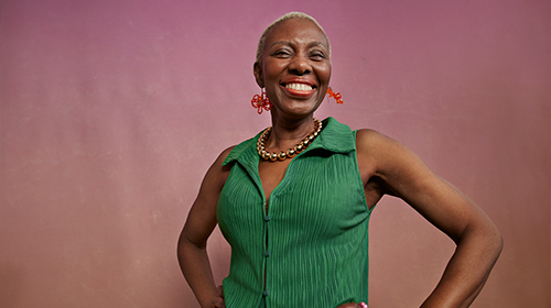 A imagem é uma foto. Uma mulher negra mais velha, de cabelos curtos e brancos, brincos vermelhos, colar dourado e blusa verde, está com as mãos na cintura e sorrindo. O fundo da imagem é rosa.