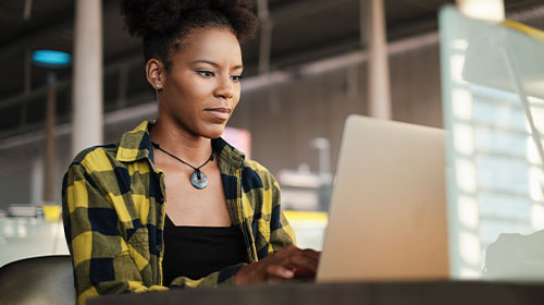 A imagem é uma foto. O foco está em uma mesa em que há um notebook virado de costas para a câmera. Mexendo no aparelho, está uma mulher negra vestindo uma camisa quadriculada nas cores amarela e preta. Ela está com o olhar focado na tela e parece estar digitando. A mulher também usa um colar com ornamento redondo pendurado no pescoço.