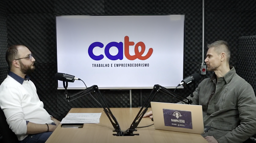 A imagem é uma foto. Em um estúdio de gravação de podcast, dois homens estão sentados, um de frente para o outro. Do lado esquerdo da imagem está um homem branco, com cabelos raspados, barba, óculos e uma blusa de frio branca. Na mesa, a sua frente, estão um microfone e uma folha de papel. Do lado direito da imagem está um homem branco, com cabelos curtos loiros e um casaco verde de frio. Na mesa, a sua frente, estão um microfone e um computador. No fundo, atrás dos dois, está um banner escrito Cate – trabalho e empreendedorismo.