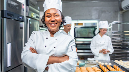 A imagem é uma foto. Em um ambiente de cozinha profissional, uma mulher negra, de cabelos curtos e crespos sorri. Ela está vestindo um uniforme branco de chef de cozinha, com chapéu e avental de botões e mangas compridas. Atrás dela estão mais duas pessoas com as mesmas vestimentas.