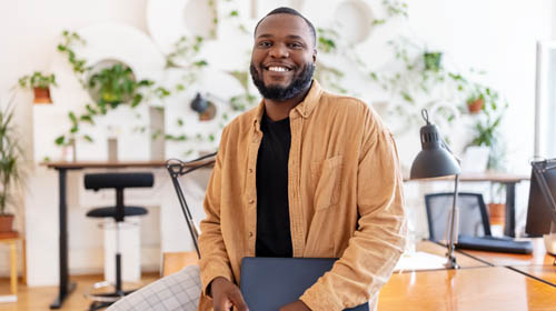 A imagem é uma foto. Um homem negro, de cabelos curtos, barba, camisa marrom e calça xadres está em um ambiente de escritório. Ele está sentado em cima de uma mesa e segura um tablet.
