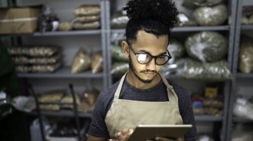A imagem é uma foto. Em um ambiente de estoque, produtos estão dispostos em prateleiras. Um homem negro, de cabelos presos, óculos, camiseta cinza e avental, está segurando uma prancheta