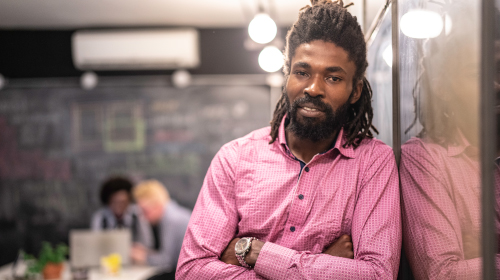 A imagem é uma foto. Em um ambiente de escritório, um homem negro, de dreadlocks e camisa social rosa, está de braços cruzados, encostado na parede.