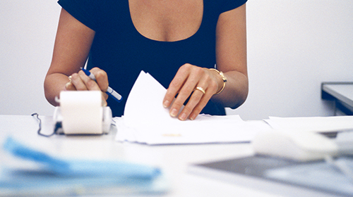 A imagem é uma foto em um espaço que parece ser um escritório. Aparece o tronco de uma mulher, sentada a uma mesa. Com a mão esquerda, ela mexe em alguns papéis que estão sobre a mesa e, com a mão direita, digita em uma máquina pequena. A mulher veste camisa azul e usa anéis e pulseiras douradas. O fundo da imagem é uma parede branca.