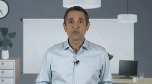 A imagem é o frame de um vídeo. Um homem com mais de sessenta anos, olha para a câmera. Ele tem cabelos curtos grisalhos e veste uma camisa social azul.