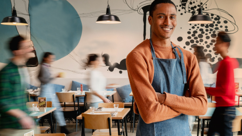 A imagem é uma foto. Em um ambiente de restaurante, com pessoas passando ao fundo, um homem de braços cruzados está em destaque. Ele é negro, de cabelos curtos, está com uma blusa de manga comprida marrom e um avental azul.