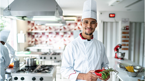 A imagem é uma foto. Em um ambiente de cozinha industrial, um homem negro está em destaque. Ele veste uniforme completo de chef de cozinha e segura uma cumbuca vermelha com temperos. Outros chefs estão ao fundo.