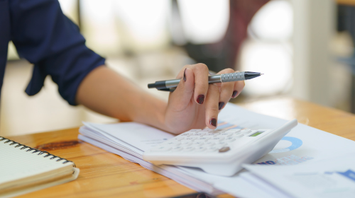 A imagem é uma foto. A mão de uma mulher branca, com unhas pintadas de marrom, segura uma caneta. Com os dedos ela toca em uma calculadora, que está apoiada em um caderno em cima de uma mesa, ao lado de outro caderno.
