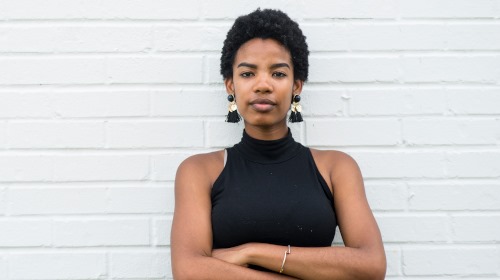 A imagem é uma foto. Ums mulher negra, de cabelos curtos, blusa e brincos pretos e está em frente a uma parede, cruzando os braços e olhando para a câmera.