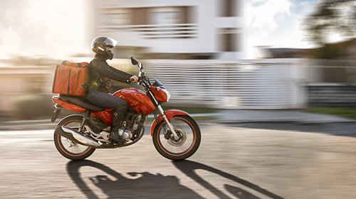 A imagem é uma foto de uma pessoa pilotando uma moto vermelha. Ela usa roupas e capacete pretos e carrega uma mala de entrega vermelha na garupa. No fundo da imagem há um prédio desfocado.