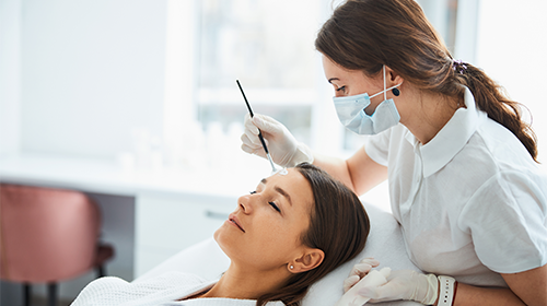A imagem é uma foto. Em um ambiente de clínica, uma profissional de estética atende uma cliente. A profissional é uma mulher branca e está paramentada com máscara e luva descartável.