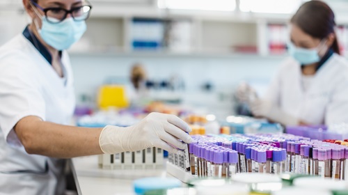 A imagem é uma foto. Em um ambiente de laboratório de análises clínicas, duas pessoas estão manuseando tubos lacrados com amostras de sangue dentro. As pessoas estão de máscara, luvas e aventais brancos.