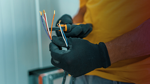 A imagem é uma foto. No foco da imagem, vemos duas mãos usando luvas pretas e segurando um conjunto de fios com as pontas desencapadas. Os fios têm cores diferentes, entre elas azul, branco, vermelho e laranja. A pessoa usa camiseta amarela e calça cinza. Não é possível ver o seu rosto.