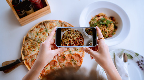 A imagem é uma foto. Em segundo plano, vemos uma mesa branca com itens gastronômicos distribuídos por sua extensão. À direita há dois pratos, um com comida e um vazio. Um pouco à esquerda, vemos uma pizza inteira e uma pequena cesta de molhos. Em primeiro plano, há duas mãos segurando um celular na horizontal e apontando a câmera para as comidas.