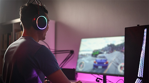 A imagem é uma foto. Encostada na parede, está uma mesa preta com um monitor, um mouse, um teclado e uma CPU dispostos sobre a sua extensão. Na cadeira de frente para a mesa, vemos um homem jogando um jogo digital com o uso do mouse e do teclado. Ele está de costas, veste uma camiseta na cor cinza e usa um fone de ouvido na cor preta.
