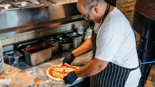 A imagem é uma foto. Nela, um pizzaiolo prepara uma pizza sobre uma bancada de granito em uma pizzaria. Ele usa óculos, veste um avental preto com listras brancas, luvas pretas e uma camiseta branca. Ao fundo, vemos ingredientes organizados em recipientes de aço inoxidável. A cena sugere um ambiente focado na produção artesanal de pizzas.