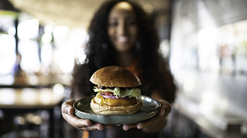 A imagem é uma foto. Em um salão bem iluminado, uma mulher de cabelos longos está de frente para a câmera com os braços estendidos enquanto segura um prato. No prato há um hambúrguer com pão, salada e queijo.