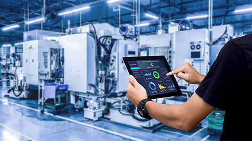 A imagem é uma foto. Em um galpão industrial, diversas máquinas na cor branca estão posicionadas ao fundo. No plano mais próximo, uma pessoa branca segura um tablet com a mão esquerda enquanto mexe na tela com a mão direita. Não é possível ver o seu rosto. A pessoa veste camiseta preta e um relógio no braço esquerdo.