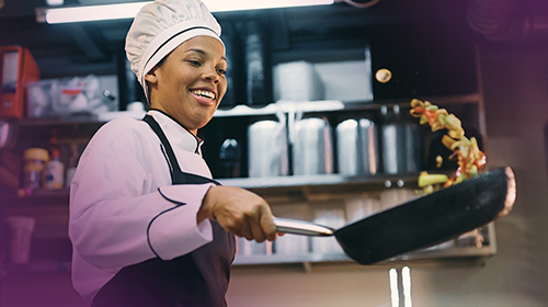 A imagem é uma foto que mostra uma chef de cozinha. Ela é uma mulher preta, veste um uniforme de chef, composto por uma touca branca, camisa branca e avental preto, está preparando um prato em uma cozinha. Ela está sorrindo enquanto salteia legumes em uma frigideira grande. O ambiente ao redor tem diversos utensílios de cozinha, como panelas, potes e equipamentos industriais.