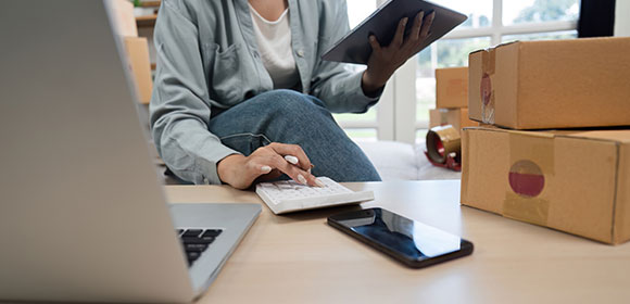 A imagem é uma foto que mostra uma pessoa sentada em um ambiente interno, vestindo uma camisa de botão cinza sobre uma camiseta branca e calça jeans. Ela segura um tablet com uma das mãos enquanto a outra mão está sobre uma calculadora branca. À sua frente, há um laptop parcialmente visível e um smartphone ao lado do teclado. Sobre a mesa, estão empilhadas diversas caixas de papelão marrom preparadas para envio, sugerindo que se trata de um empreendedor ou vendedor online organizando pedidos.