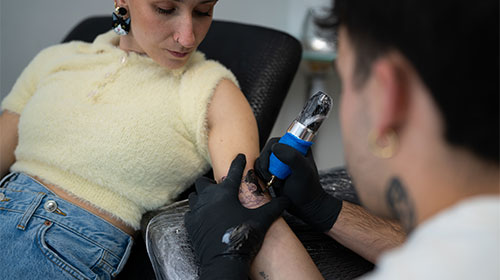A imagem é uma foto. Em um estúdio de tatuagem, uma mulher branca está sentada em uma cadeira reclinável preta. Ela veste uma blusa de lã branca e calça jeans. O seu braço esquerdo está esticado, enquanto uma pessoa apoia uma máquina de tatuagem sobre ele. O rosto do tatuador não aparece. Ele usa luvas pretas e segura uma máquina de tatuagem.