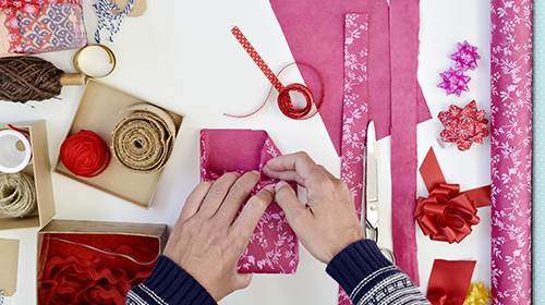 A imagem mostra as mãos de uma pessoa embrulhando uma pequena caixa com papel de presente rosa estampado. As mãos estão dobrando o papel ao redor da caixa. A cena está sobre uma mesa branca, repleta de materiais para embalagem e decoração de presentes, como fitas vermelhas e douradas, rolos de barbante, tesoura, papel de seda rosa, laços decorativos e outras pequenas caixas de papelão abertas com fitas dentro.