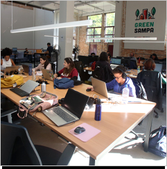 A imagem &eacute; uma foto. Em uma sala ampla e iluminada, h&aacute; diversas mesas de trabalho colaborativo com notebooks, garradas de &aacute;gua e outros itens de uso profissional. Ao fundo, uma placa branca pendurada na parede cont&eacute;m a express&atilde;o Green Sampa.