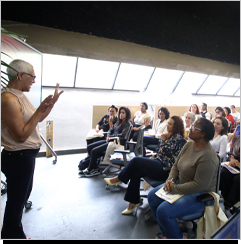 Em uma sala ampla de janelas grandes e teto preto, diversas mulheres est&atilde;o sentadas em fileiras de cadeiras de escrit&oacute;rio. Elas olham para a frente, na dire&ccedil;&atilde;o de uma mulher de cabelo grisalho e curto, que est&aacute; de p&eacute;.