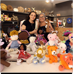 A imagem &eacute; uma foto. Em uma sala de paredes pretas, h&aacute; um espelho &agrave; esquerda. Na parede, h&aacute; prateleiras com diversos itens de artesanato. O foco est&aacute; em duas mulheres que seguram itens de artesanato enquanto posam para a c&acirc;mera, sorrindo. &Agrave; frente delas, h&aacute; uma mesa com diversos itens como bonecas e figuras de croch&ecirc;.