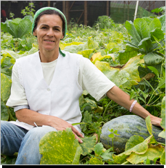 A imagem &eacute; uma foto. H&aacute; uma planta&ccedil;&atilde;o baixa com diversas folhas em tons verdes e amarelados. O foco est&aacute; em uma mulher branca sentada nomeio das folhas. Ela veste jaleco branco e cal&ccedil;a jeans, apoia os bra&ccedil;os para a sua esquerda, segurando um grande vegetal da planta&ccedil;&atilde;o.