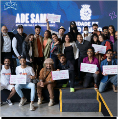 A imagem &eacute; uma foto. Ao fundo h&aacute; um painel azul com o logo da prefeitura de S&atilde;o Paulo. Em primeiro plano, diversas pessoas posam para a c&acirc;mera, sorrindo. Algumas delas seguram pap&eacute;is com as duas m&atilde;os