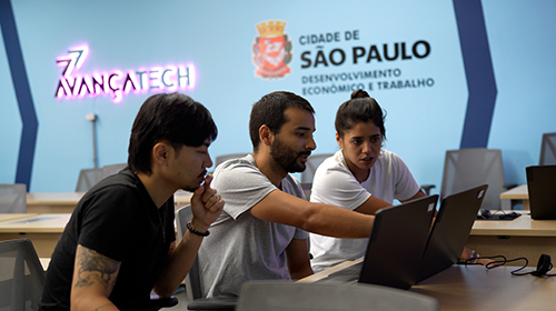 A imagem é uma foto. Ao fundo, uma parede azul apresenta o símbolo do Avança Tech e o símbolo da prefeitura de São Paulo. No plano mais próximo, três pessoas estão sentadas lado a lado. Elas olham para um monitor com um olhar atento, enquanto a pessoa do meio estende a mão apontando para a tela.