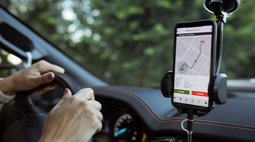 A imagem é uma foto. O ambiente é o interior de um carro. O foco está no painel de um carro, onde há um suporte para celular. O aparelho que está no suporte mostra uma tela de mapa com um trajeto delineado e dois botões abaixo, um verde e um vermelho. À esquerda do celular, vemos duas mãos de uma pessoa branca sobre o volante do carro.