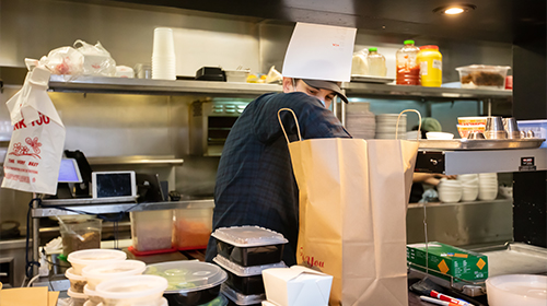 A imagem é uma foto. Nela, vemos um trabalhador em uma cozinha comercial preparando pedidos para entrega. O homem está atrás de uma bancada, ele usa máscara de proteção e está com as mãos dentro de um saco de papel para delivery, sugerindo que está colocando uma comida na embalagem. Na bancada, é possível observar vários recipientes plásticos transparentes com alimentos preparados. Atrás do homem, há embalagens vazias empilhadas, molhos em grandes recipientes plásticos e um forno elétrico.