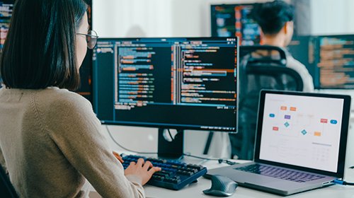 A imagem é uma foto de um ambiente de trabalho moderno com foco em tecnologia e programação. Em primeiro plano, vemos uma mulher de cabelos lisos e escuros, na altura dos ombros, usando óculos e um suéter claro. Ela está sentada de costas para a câmera, digitando em um teclado posicionado à frente de dois monitores grandes. As telas mostram códigos coloridos em fundo escuro, sugerindo que ela está programando. À sua direita, há um notebook com a tela aberta exibindo um fluxograma ou diagrama de processos com blocos coloridos. Ao fundo, desfocado, está outro profissional também sentado e olhando para um monitor com códigos.