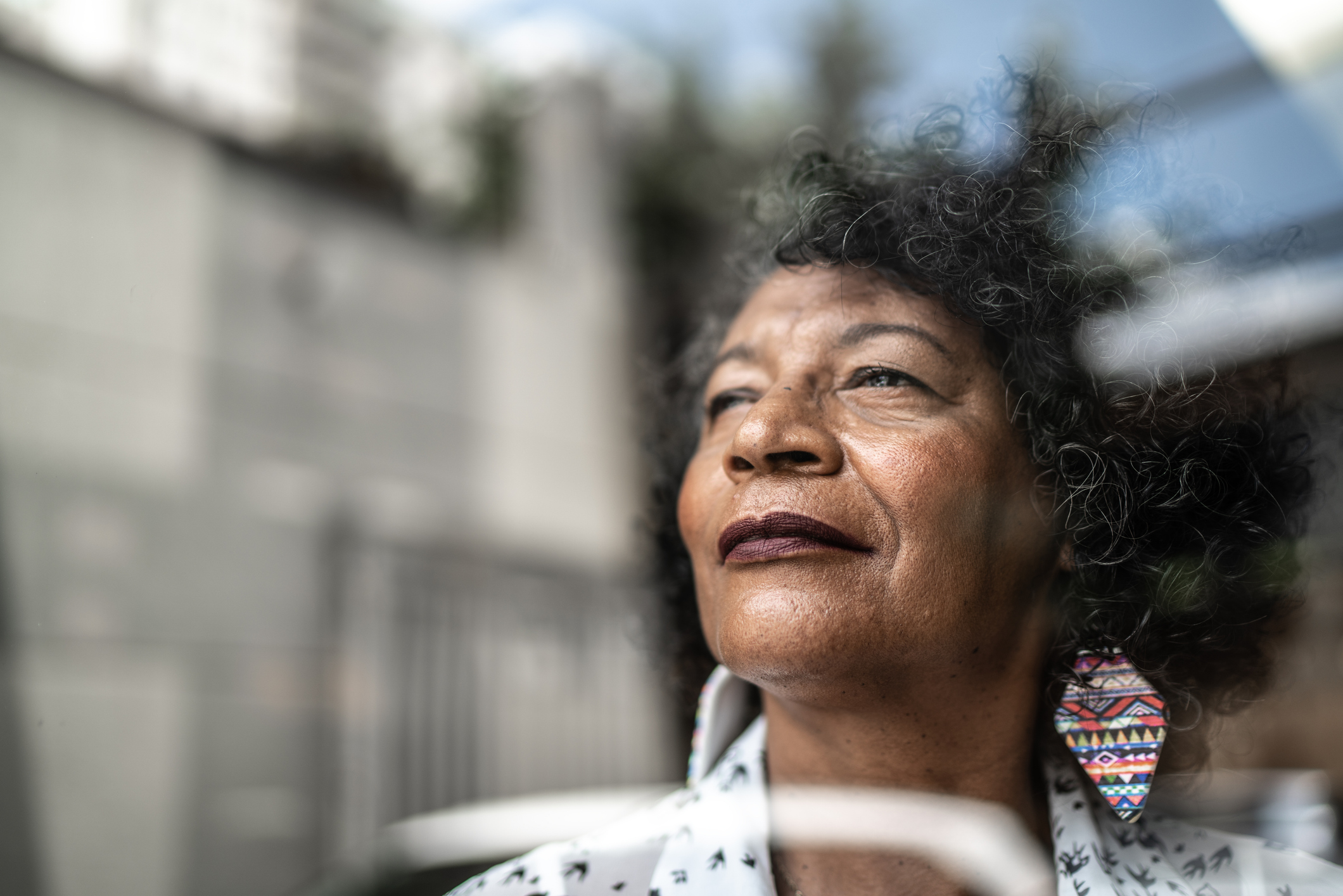 A imagem é uma foto. O foco está em uma mulher idosa. Ela é negra e tem cabelos em tons castanho-escuros e grisalhos. Ela usa batom roxo, colares coloridos em formato de losango e, no momento da foto, olha para o horizonte através de um vidro.