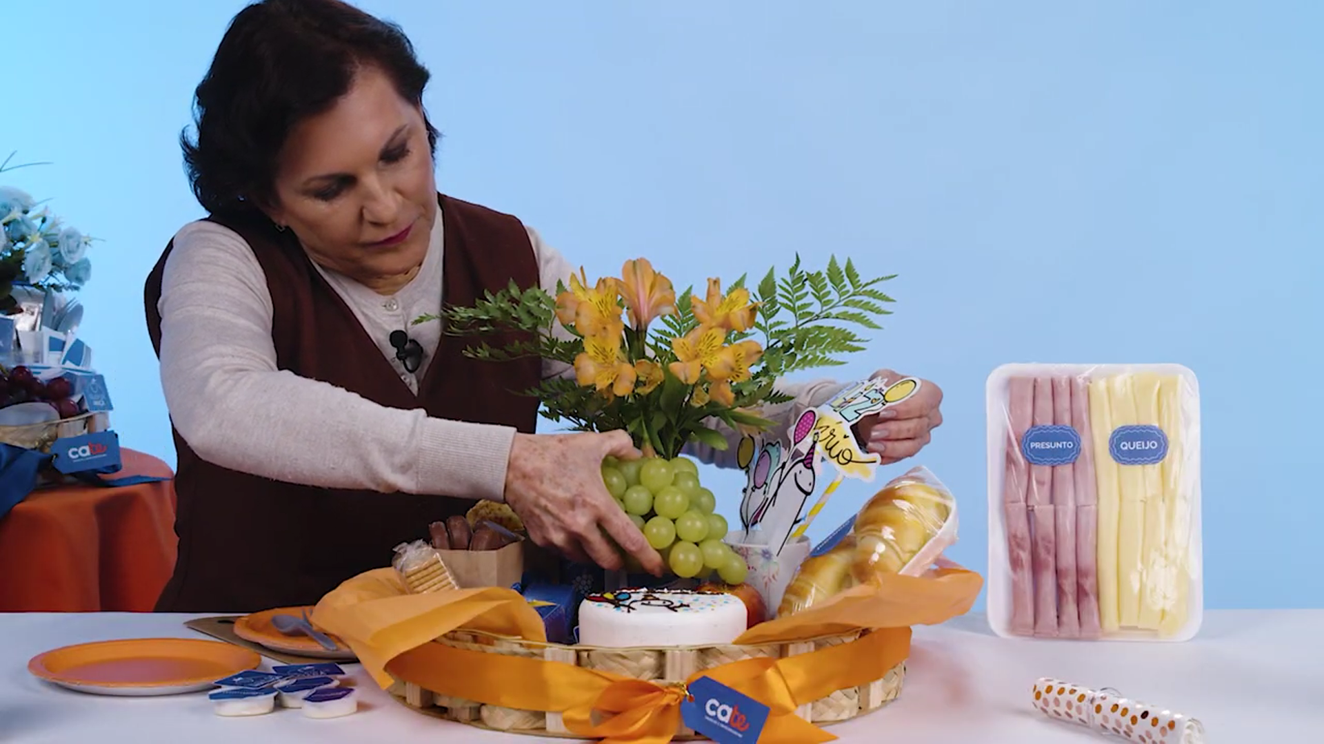 Uma mulher monta uma cesta de café da manhã decorada com laço laranja. Dentro da cesta há flores amarelas, uvas, pães, biscoitos e bolo. Ao lado, sobre a mesa, estão frios fatiados (presunto e queijo) e pratos. O fundo é azul claro.